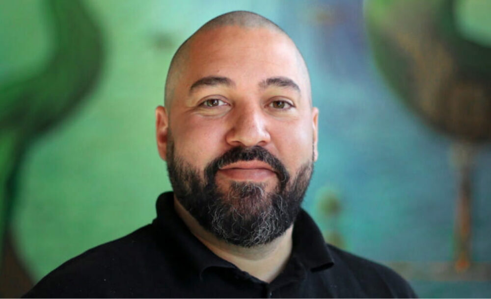 Porträtt på en man med rakat hår och mörkt skägg mot en grön bakgrund.