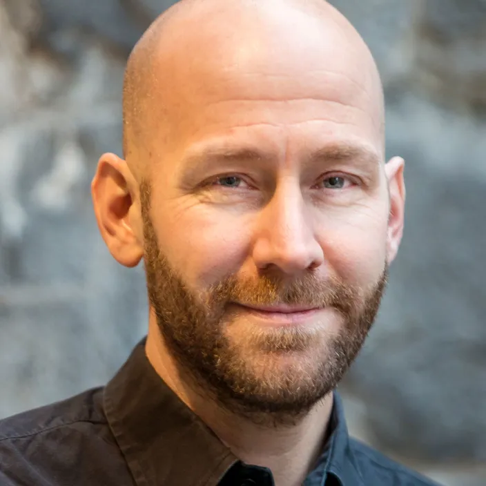 Porträtt på en man med mörk skjorta och skägg. Inget hår p huvudet. 