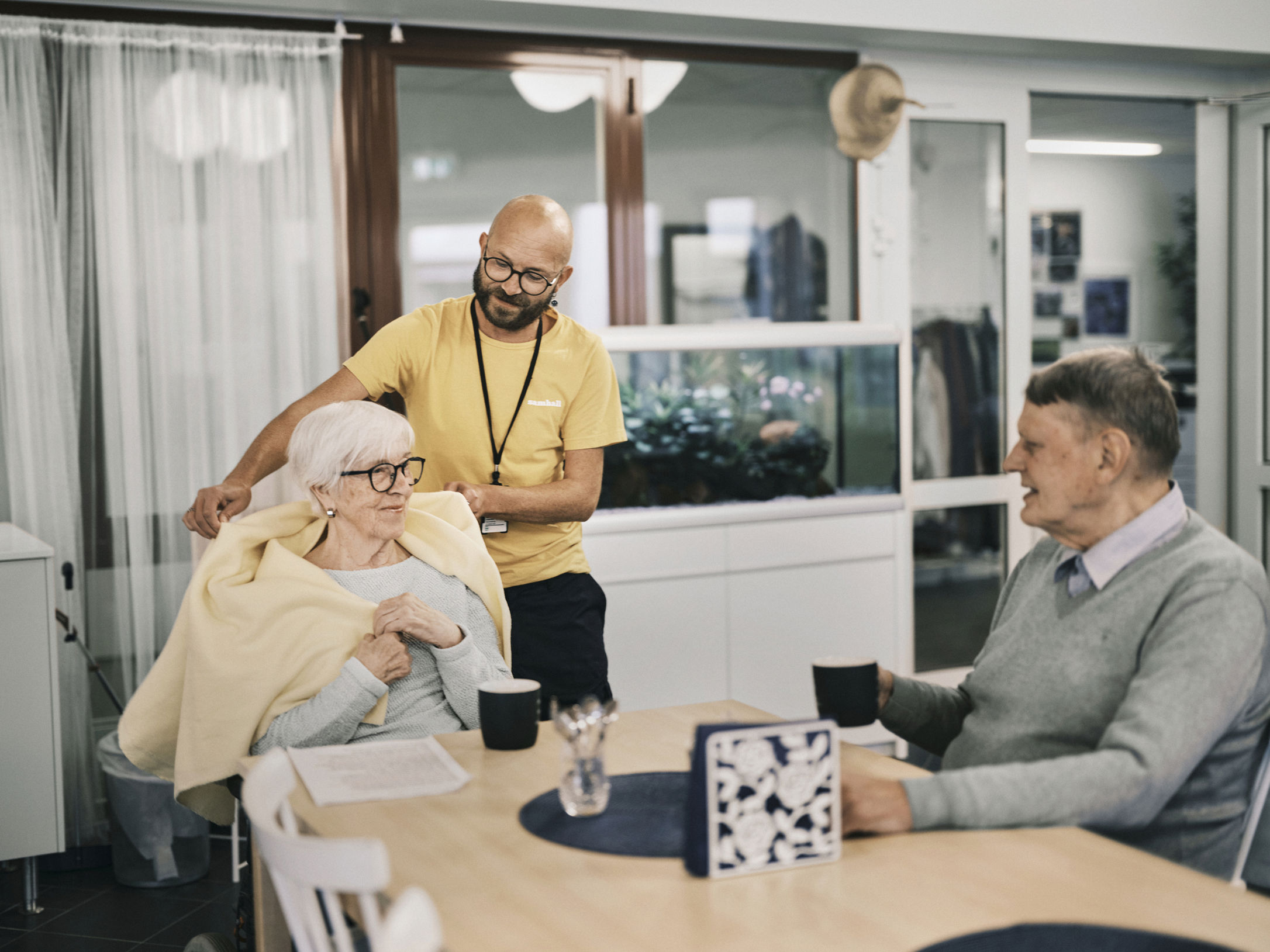 En Samhallmedarbetare interagerar med två äldre personer vid ett matbord