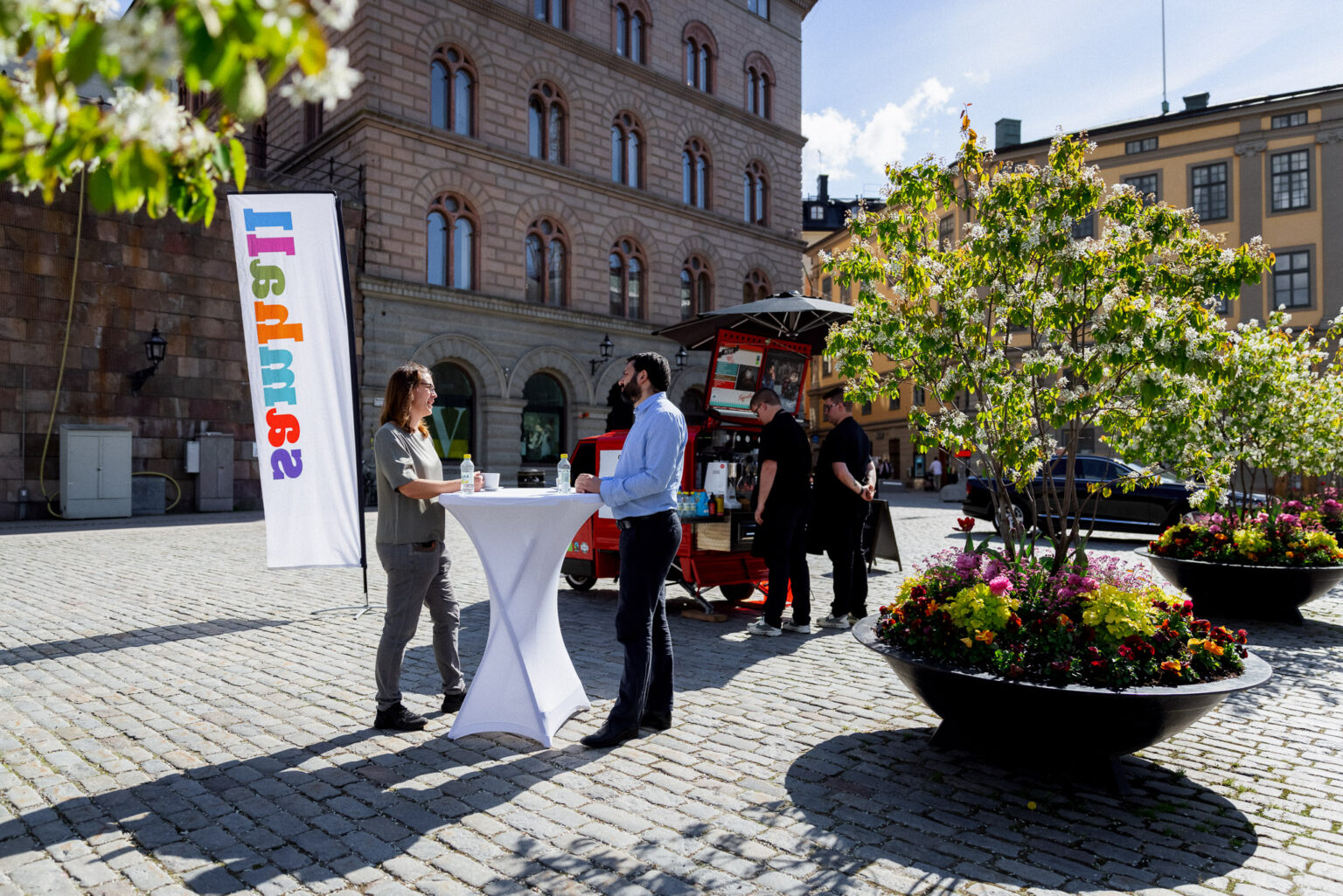 Personer på ett torg runt ett bord samt en kaffevagn