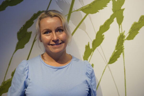 Portätt av Jenny Lindstrand som står framför en fondvägg med gröna blad. 