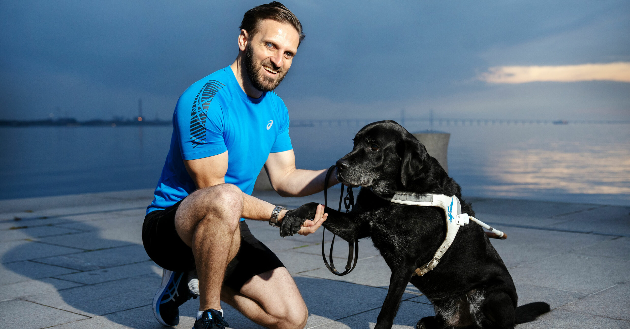 Fatmir sitter på huk på en brygga vid vattnet och håller i sin svarta hunds ena tass.