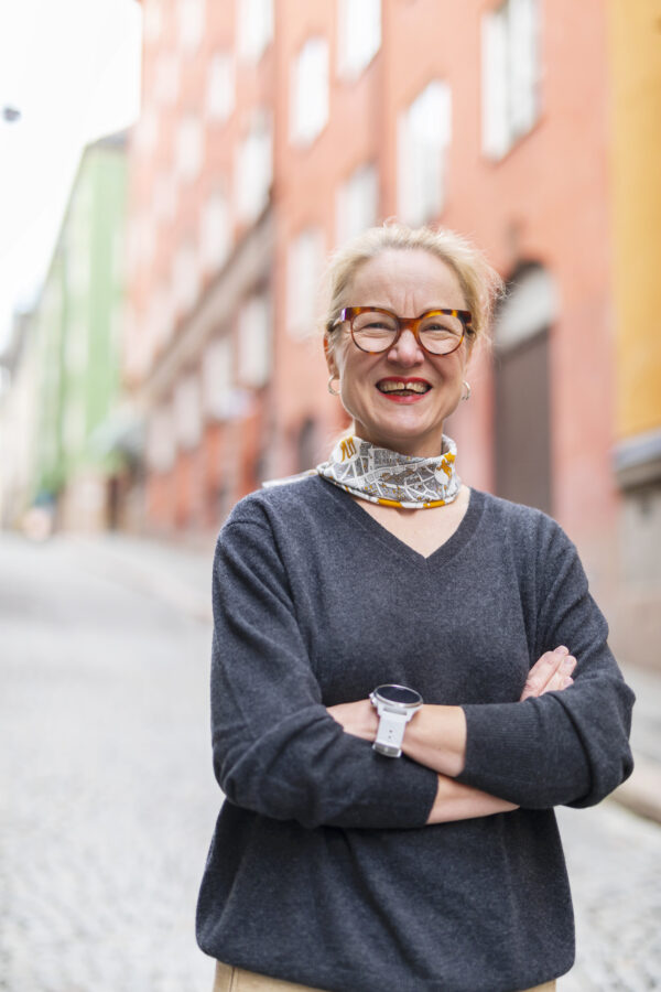 Ulrika Årehed Kågström ler in i kameran och har armarna i kors. Hon står framför blurrade lägenhetshus. 