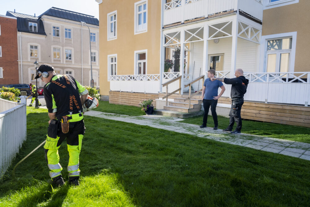 Det är sol på bilden. Samhall-medarbetaren Michael Johnson trimmar gräskanten framför ett hus, medan Samhalls områdeschef Martin Lärnestad och Niklas Eklund,, fastighetsskötare på Heimstaden, inspekterar området.