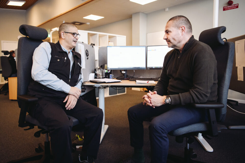Två personer sitter i kontorsstolar framför ett skrivbord och pratar.