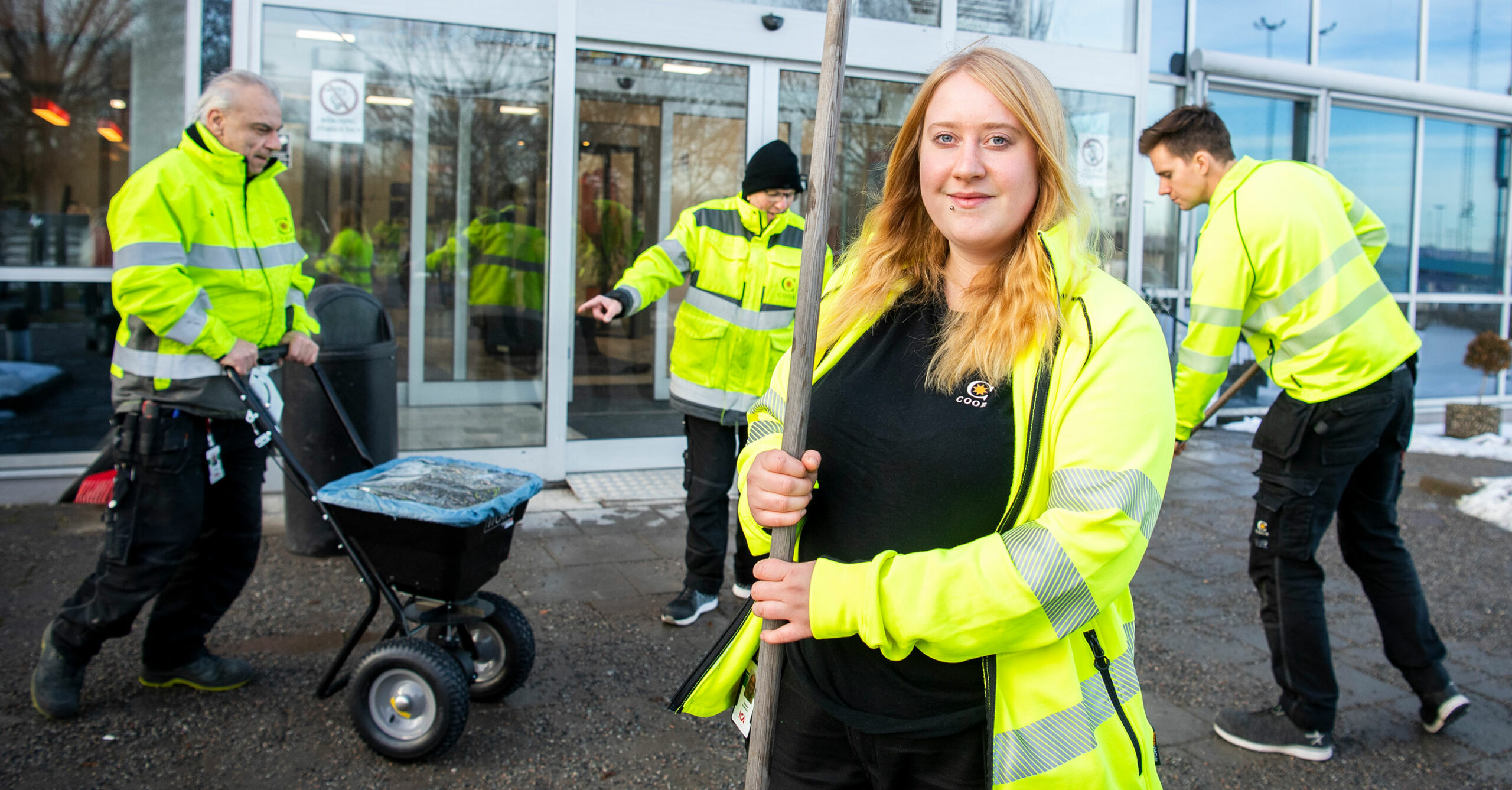 Medarbetare utomhus ståendes vid en byggnads entré. De bär reflexjackor och håller i verktyg för att lägga ut grupp på marken.