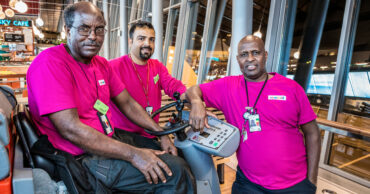 Bild på tre medarbetare bärandes lila tröjor på Arlanda. En av personerna sitter i en maskin som putsar golvet.