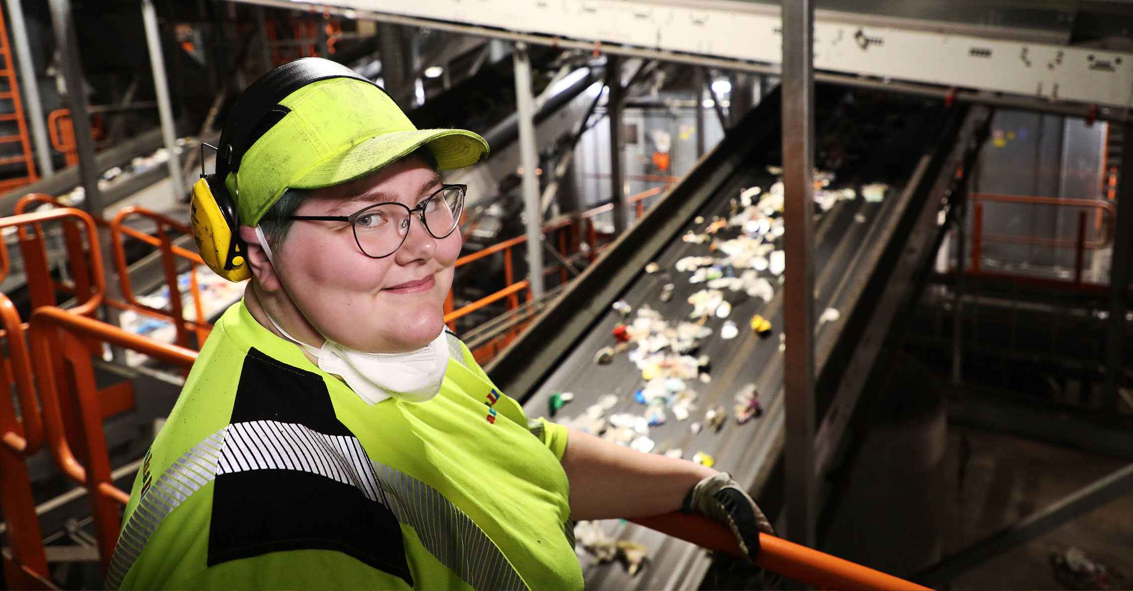 Porträtt av Jennifer Sjöberg som tittar in i kameran. Hon befinner sig framför ett rullband som fraktar plaståtervunnet material.