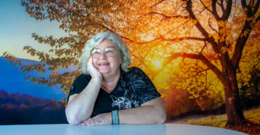 Porträtt av Gudrun som sitter lutad mot ett bord. Bakgrund är ett höstmotiv med gula löv på ett träd. Hon har grått lockigt hår.
