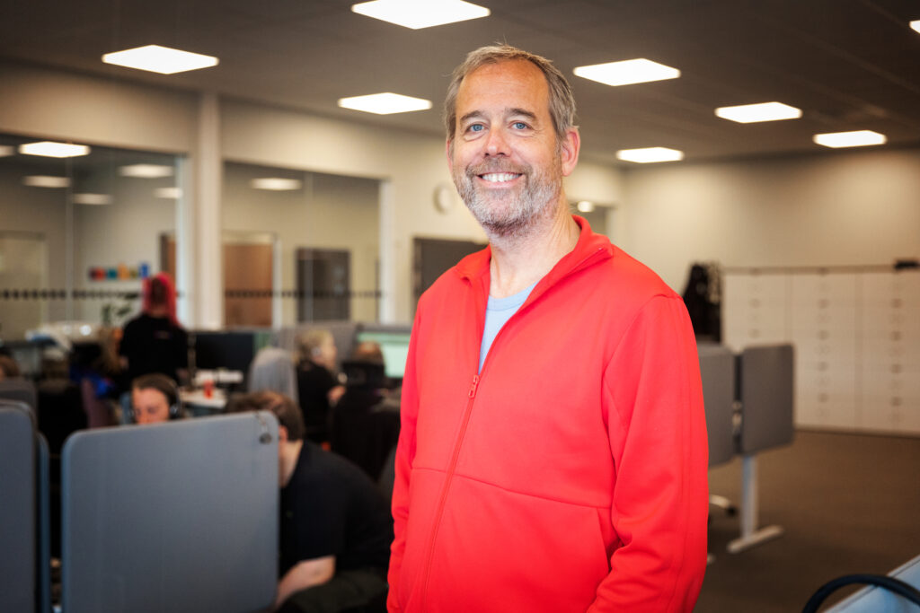 Jörgen ler och tittar in i kameran. Han bär en röd tröja och står i ett öppet kontorslandskap. 