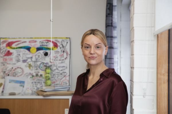 Porträtt av Alma Moslan som står i ett kontor och tittar in i kameran. I bakgrunden syns en färgglad illustration. 