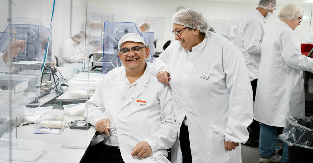 Två medarbetare på Beneli skrattar på jobbet. De bär vita rockar och skyddsnät på huvudet.