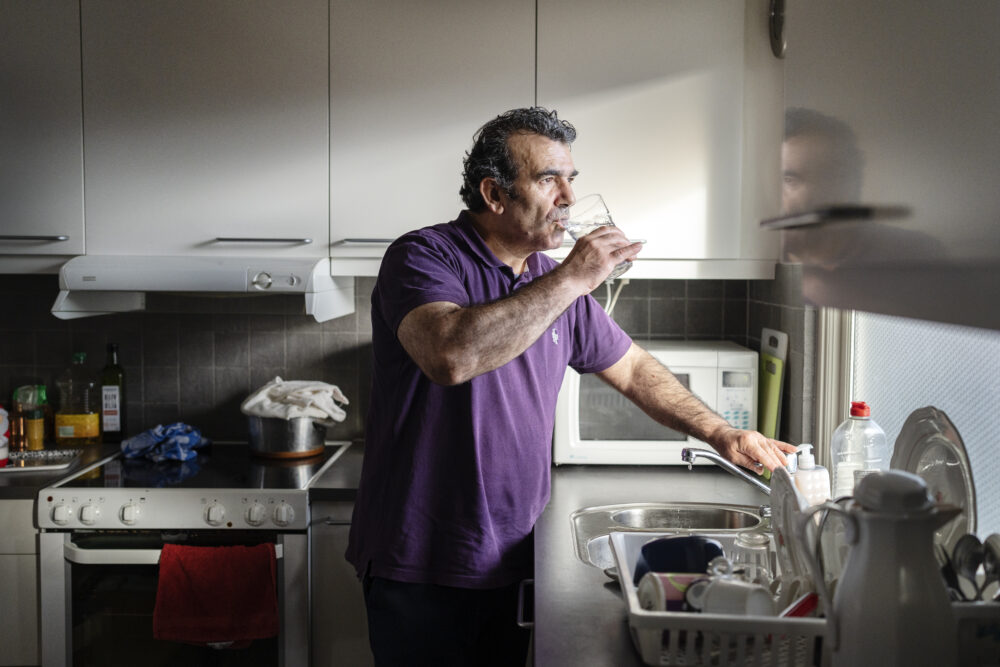 Porträtt på Parvis som dricker ett glas vatten i ett kök. Han bär en lila. pikétröja och tittar ut genom ett fönster. 