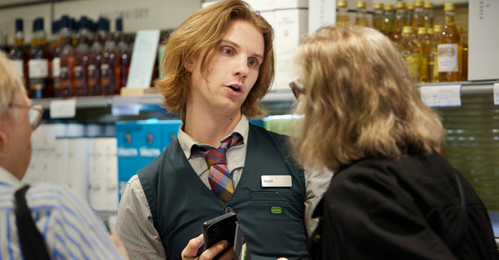 Bild på Johan som hjälper två kunder i systembolagets butik.