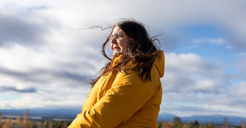 Bild på en kvinna som står utomhus med en gul jacka. Vinden gör att hennes mörka hår fladdrar till.