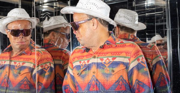 Bild på en man som bär en cowboy-hatt. Han har en färgglad skjorta och står vid en spegelvägg.